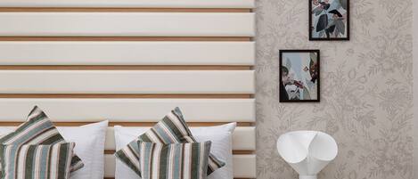 A bedroom with a striped bedspread, white pillows, a wooden headboard, a white nightstand with a lamp, and floral wallpaper.