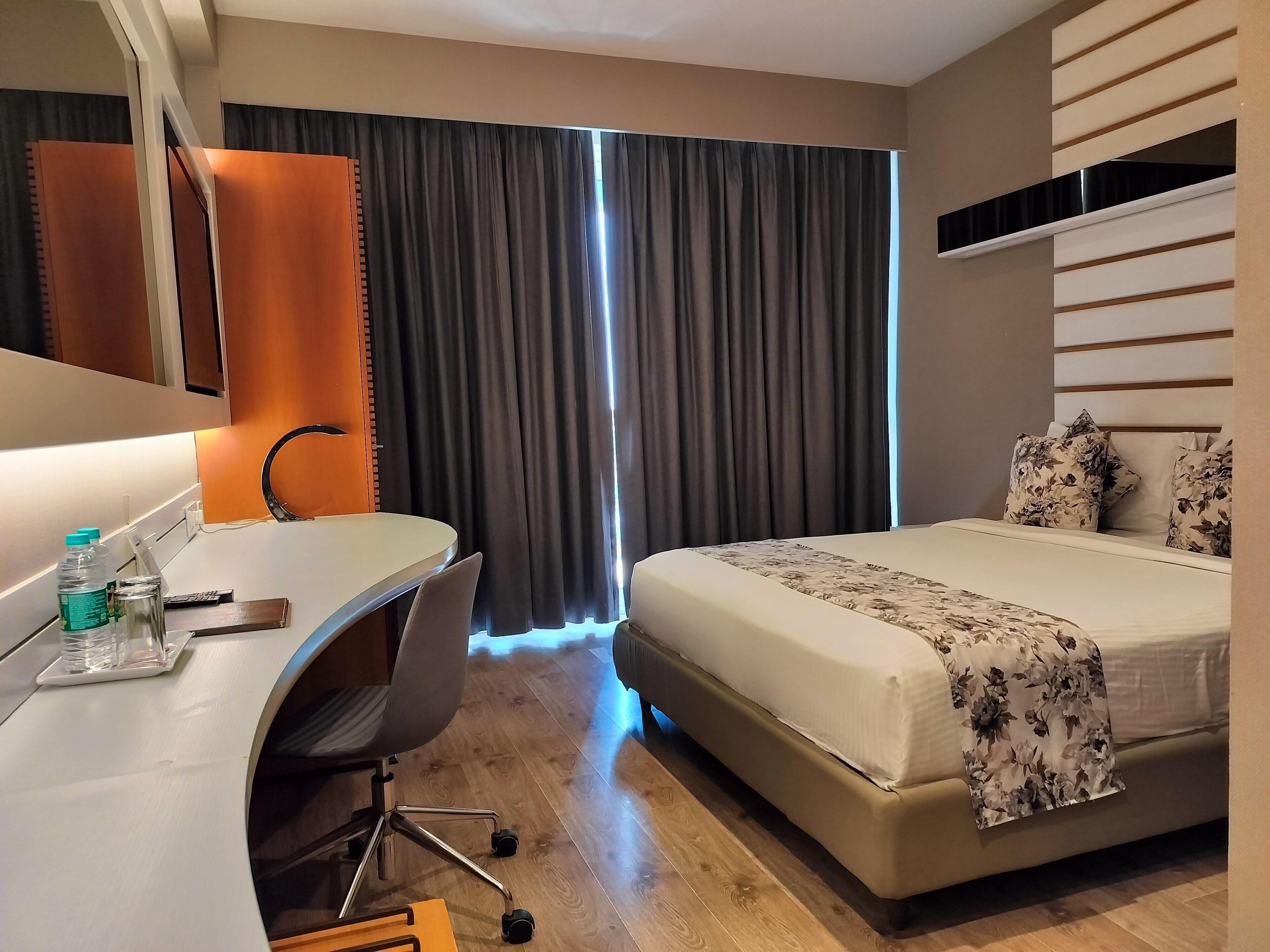 Une chambre d’hôtel avec un lit, un bureau et une chaise. Il y a une fenêtre avec des rideaux, un miroir et une étagère fixée au mur.