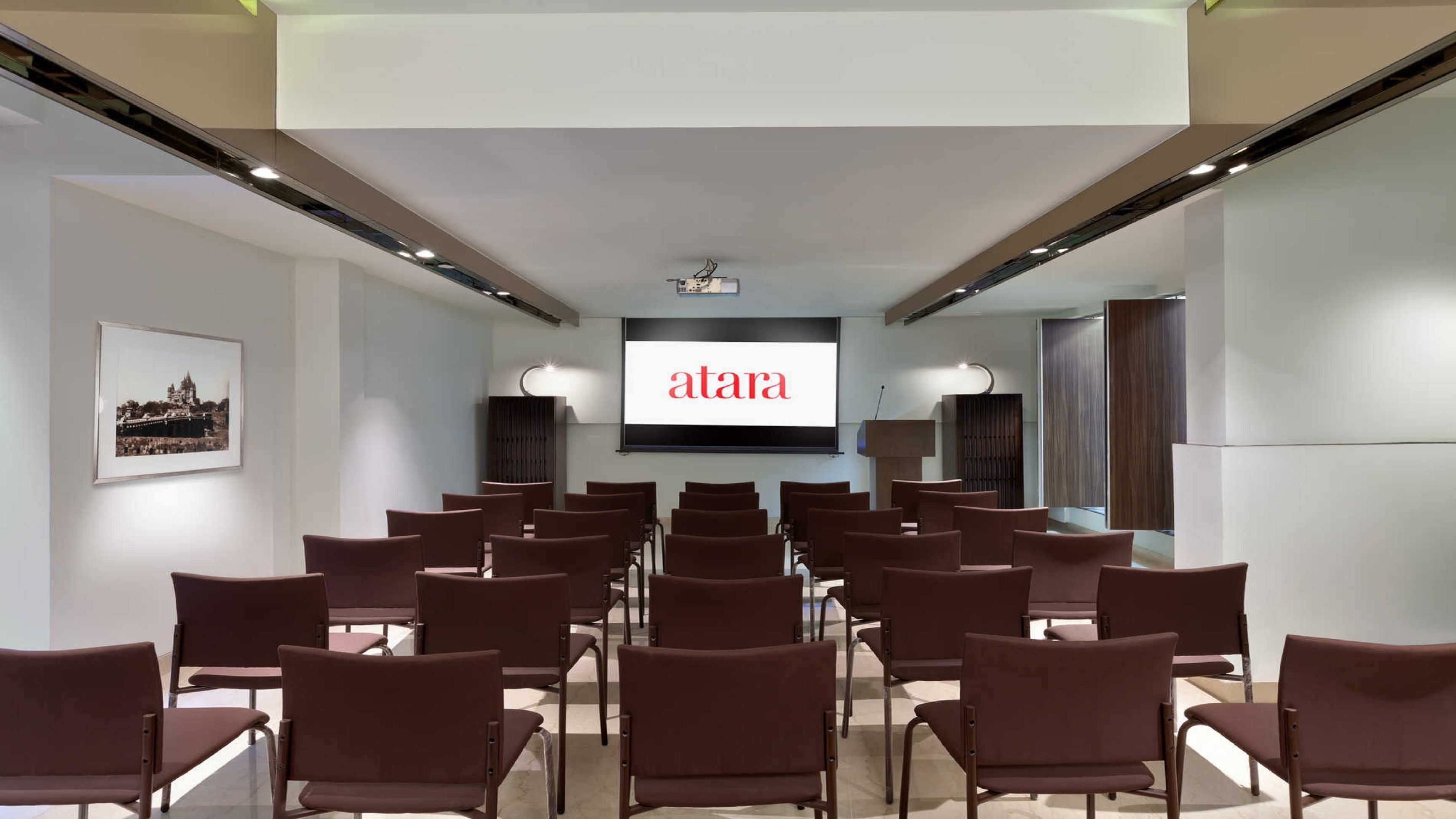 A conference room with rows of brown chairs facing a large screen displaying "atar."
