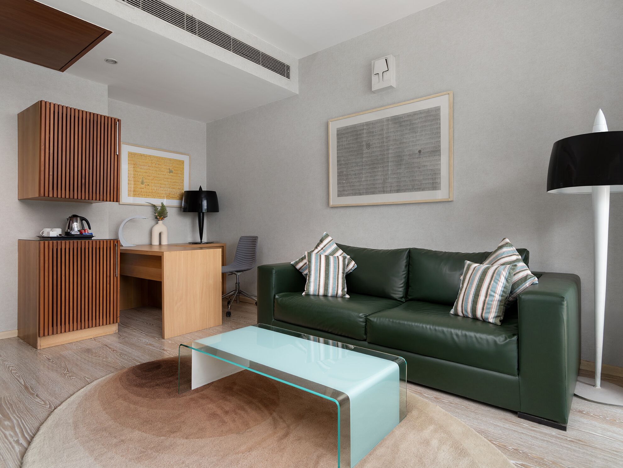 A modern living room with a green leather sofa, a glass coffee table, a wooden desk, and a framed artwork on the wall.