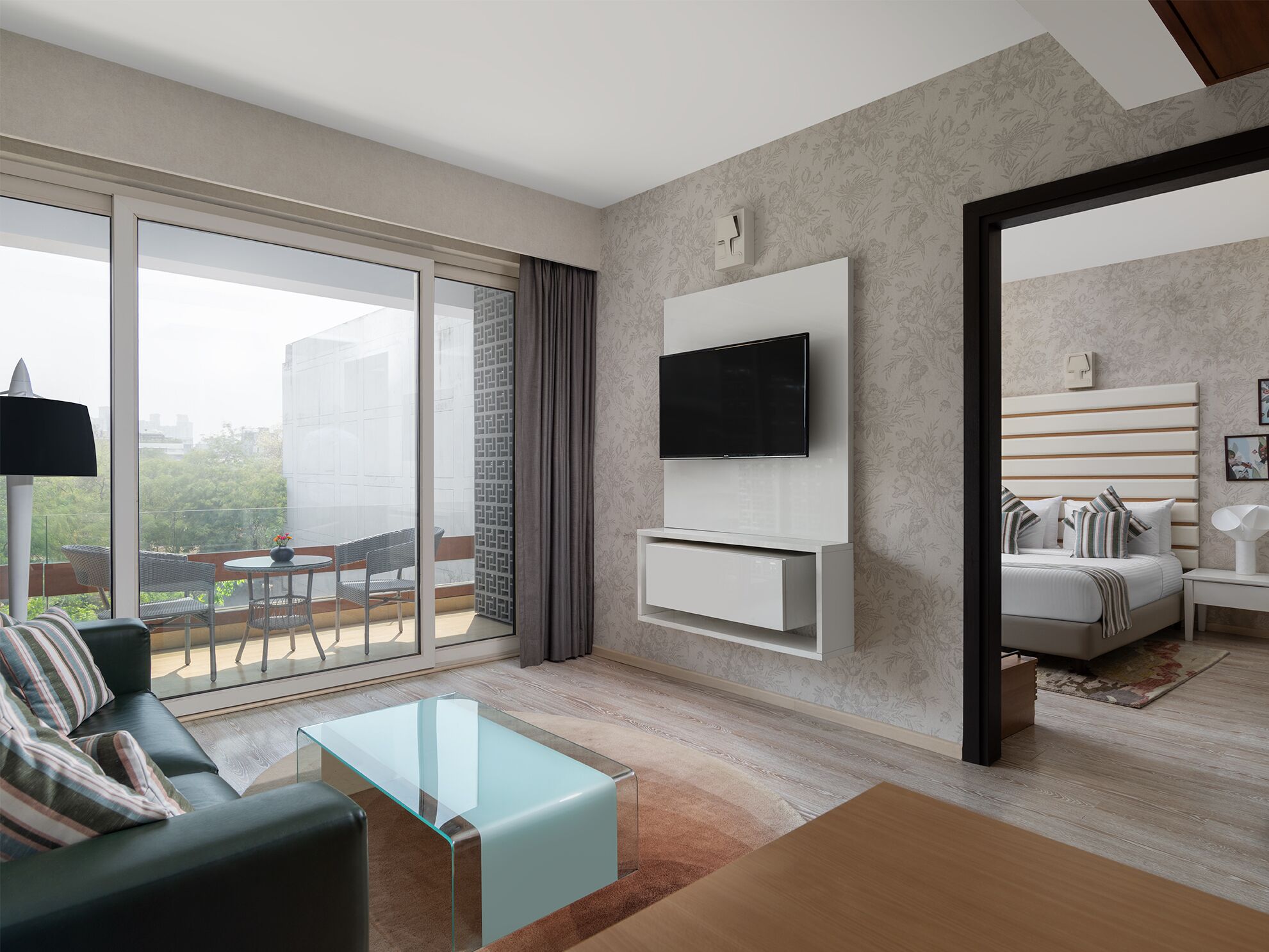 A modern living room with a large TV, a sofa, a glass coffee table, and a balcony with outdoor furniture.