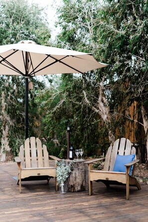 Two wooden Adirondack chairs with cushions, a table with a vase and wine glasses, and a large umbrella. - Saltbush Retreat (Longreach)