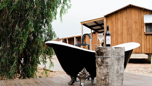 Une terrasse en bois avec une baignoire noire, un seau en bois et une bouteille de vin.