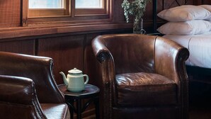 Une pièce avec une grande fenêtre, un lit avec des oreillers, un fauteuil en cuir, une petite table avec une théière et des appliques murales.