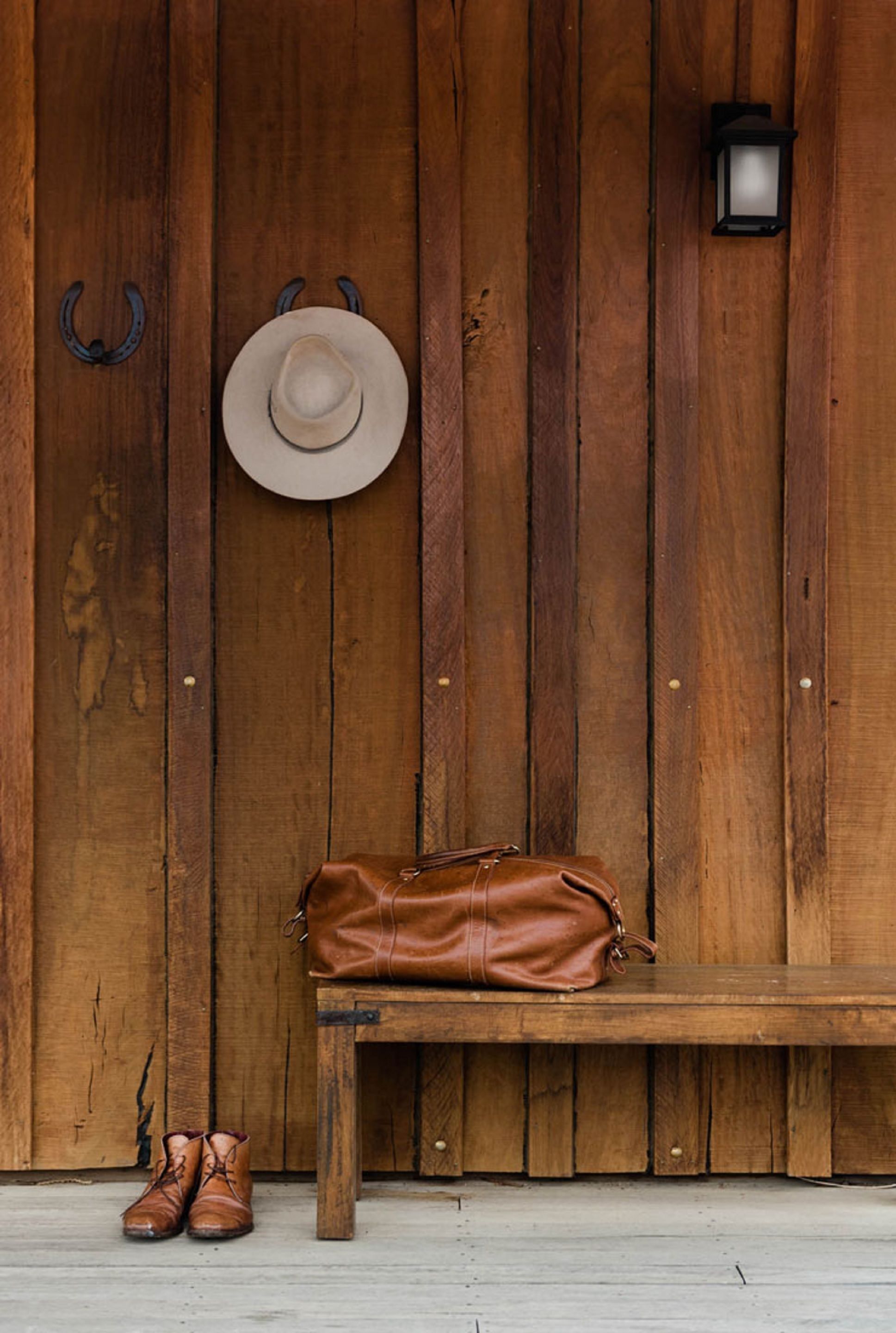Un banco de madera con un bolso de cuero marrón y un sombrero colgado en la pared.