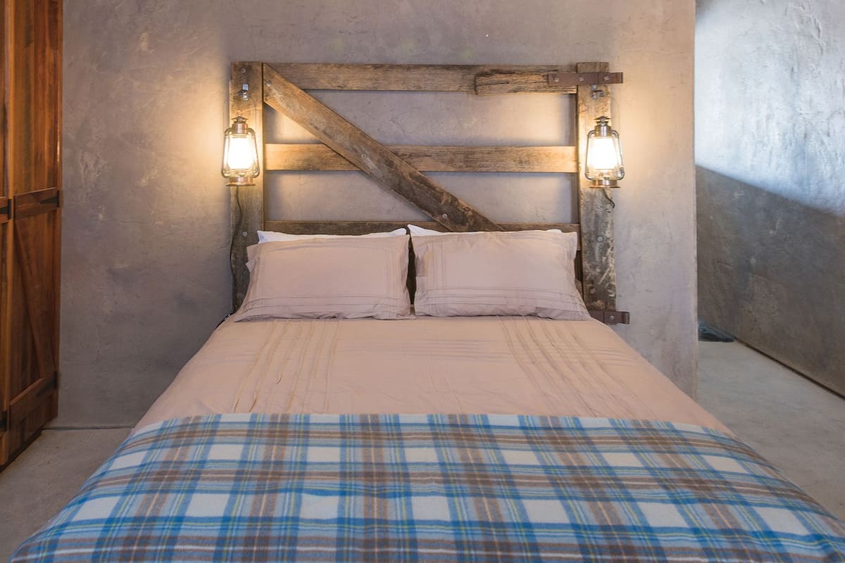 A bed with a plaid bedspread, a wooden headboard with two wall-mounted lights, and a wooden wardrobe to the left.