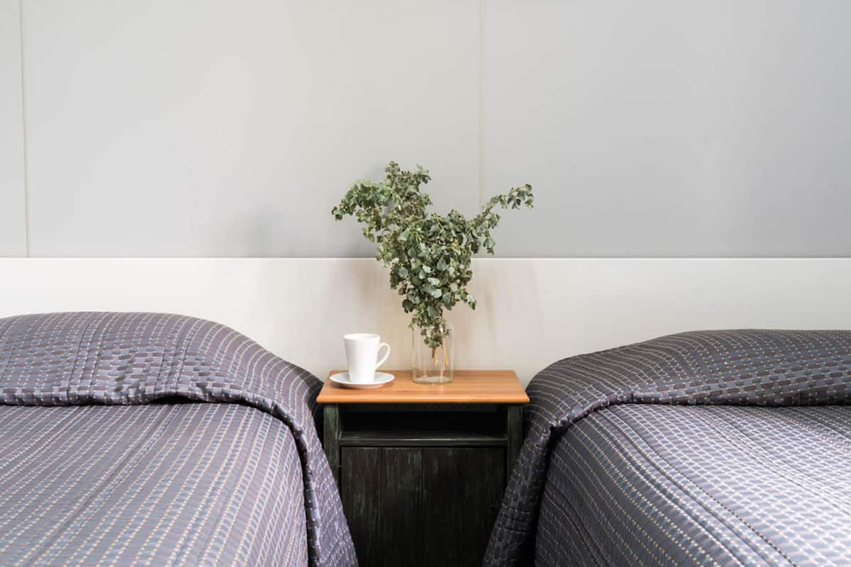 A hotel room with two beds, a small wooden nightstand, a white cup, and a vase with a plant.