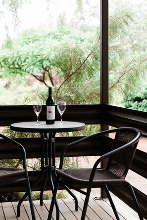 A round table with two chairs, a bottle of wine, and two glasses on a balcony with a view of greenery. - Saltbush Retreat (Longreach)
