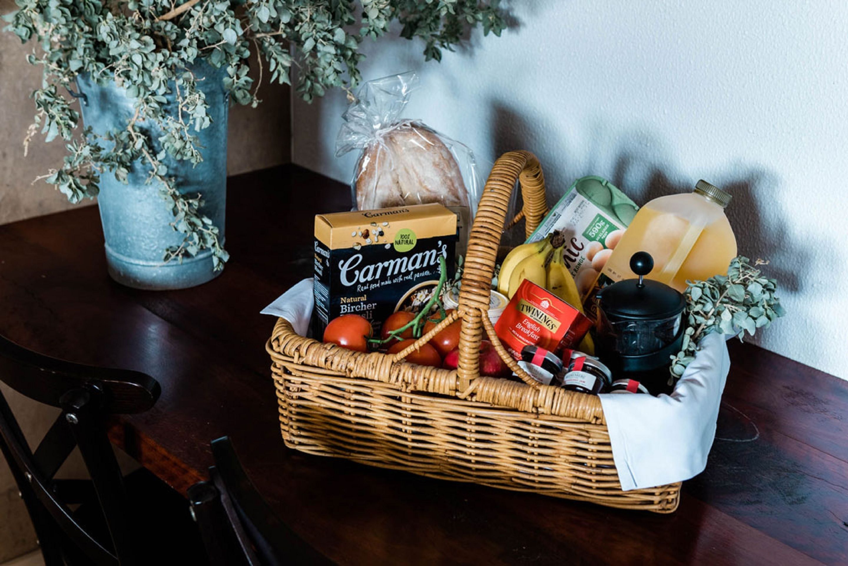 A wicker basket filled with groceries, including bread, tomatoes, and a bottle of juice, on a wooden table.