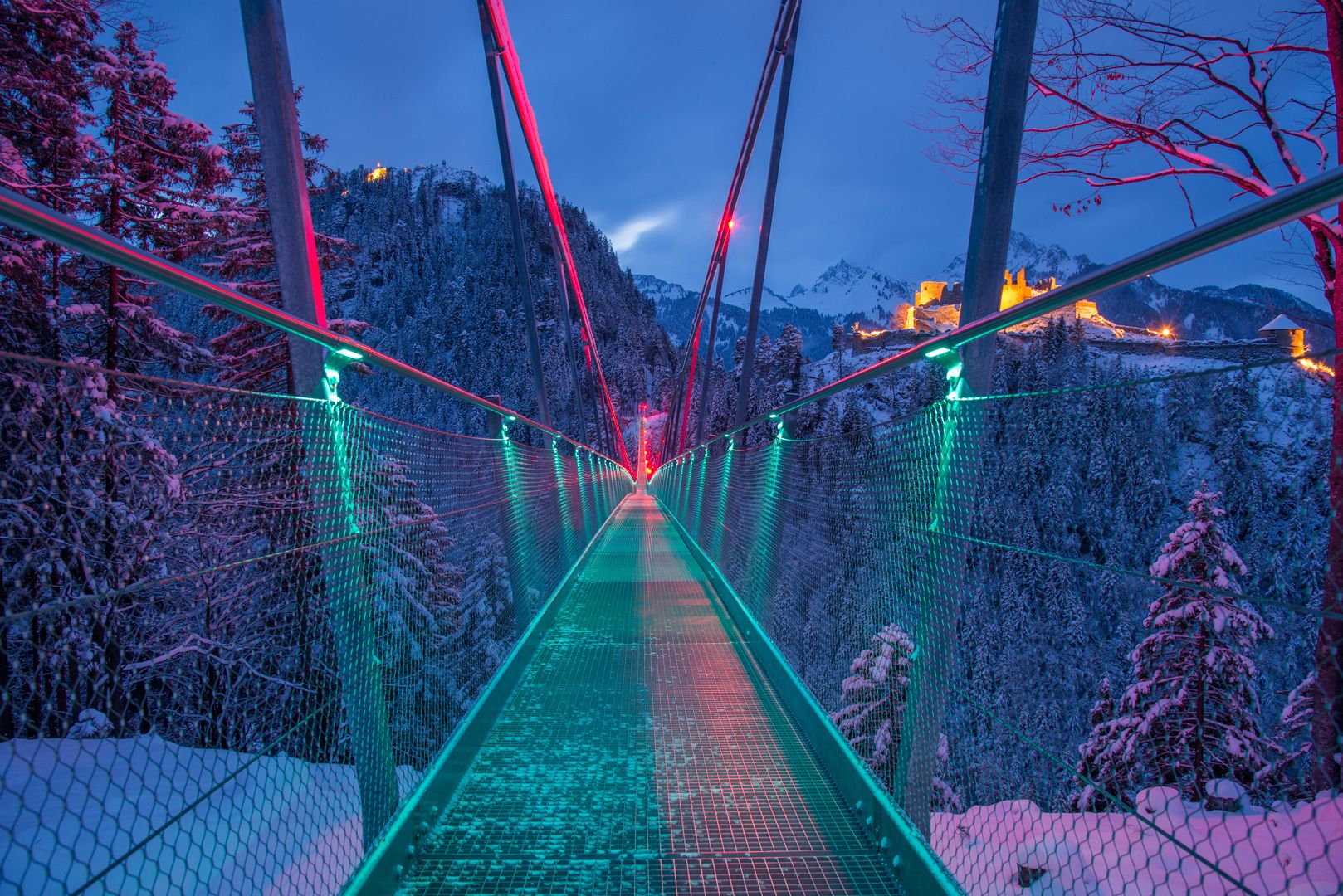 A suspension bridge with a metal walkway, surrounded by snow-covered trees and mountains, illuminated by red and green lights.