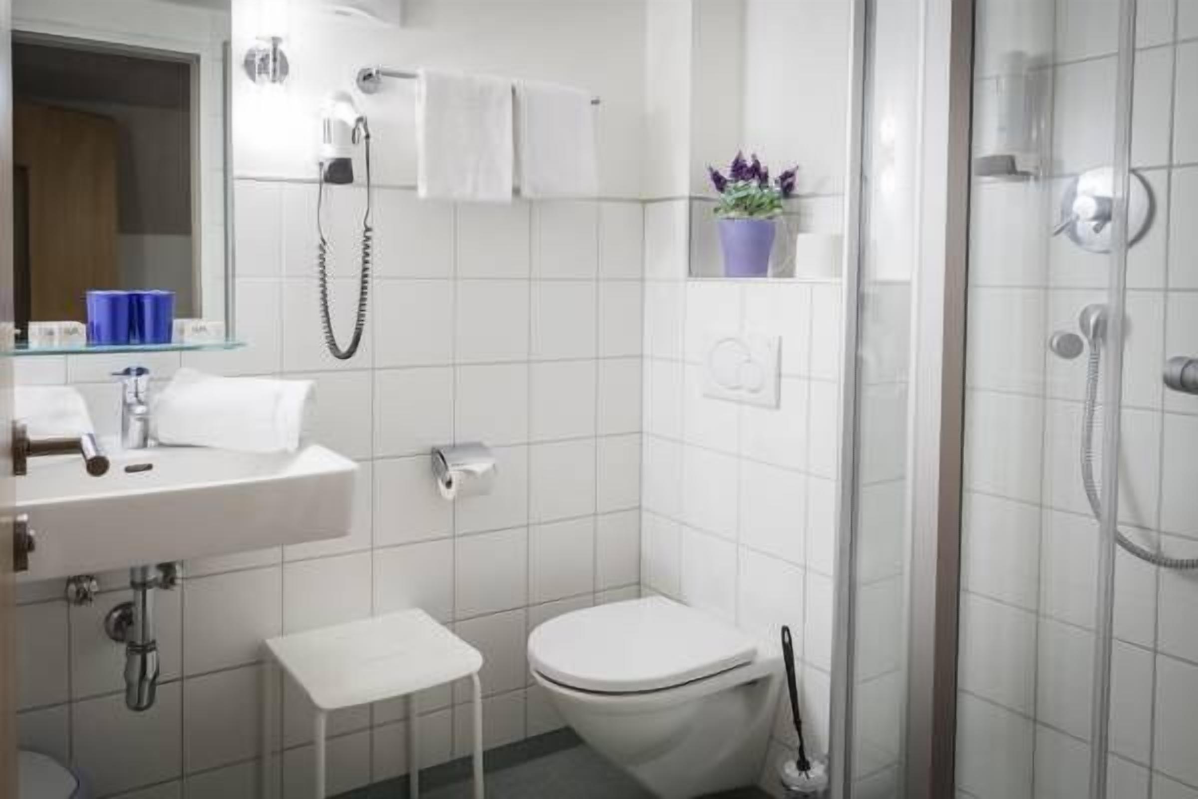 Ein modernes Badezimmer mit Waschbecken, Toilette und Dusche in Weiß. Auf dem Waschtisch steht ein kleiner weißer Hocker und ein Blumentopf.
