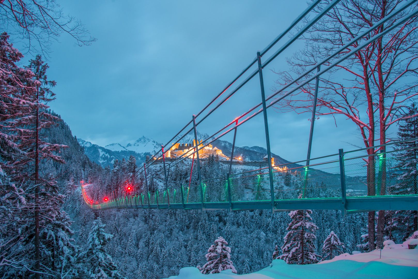 A snow-covered landscape with a suspension bridge and a mountain in the background.