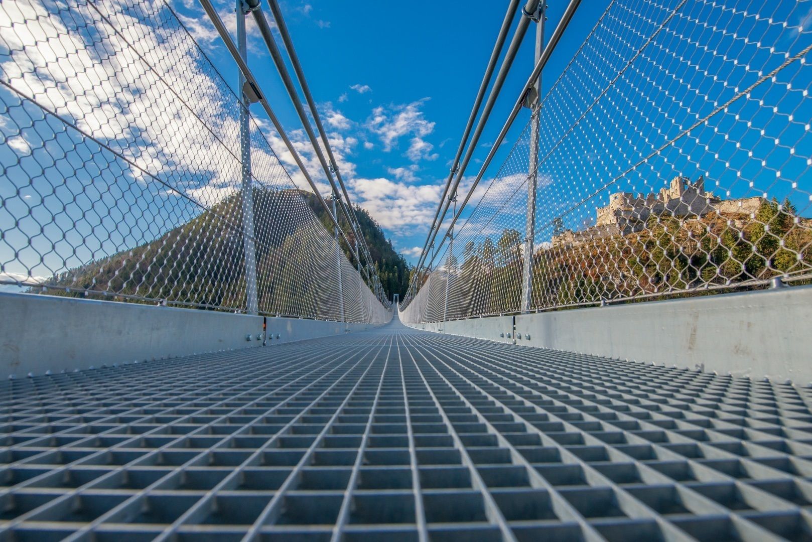 A pedestrian bridge with a metal grid floor and chain-link safety netting, leading to a mountainous landscape.