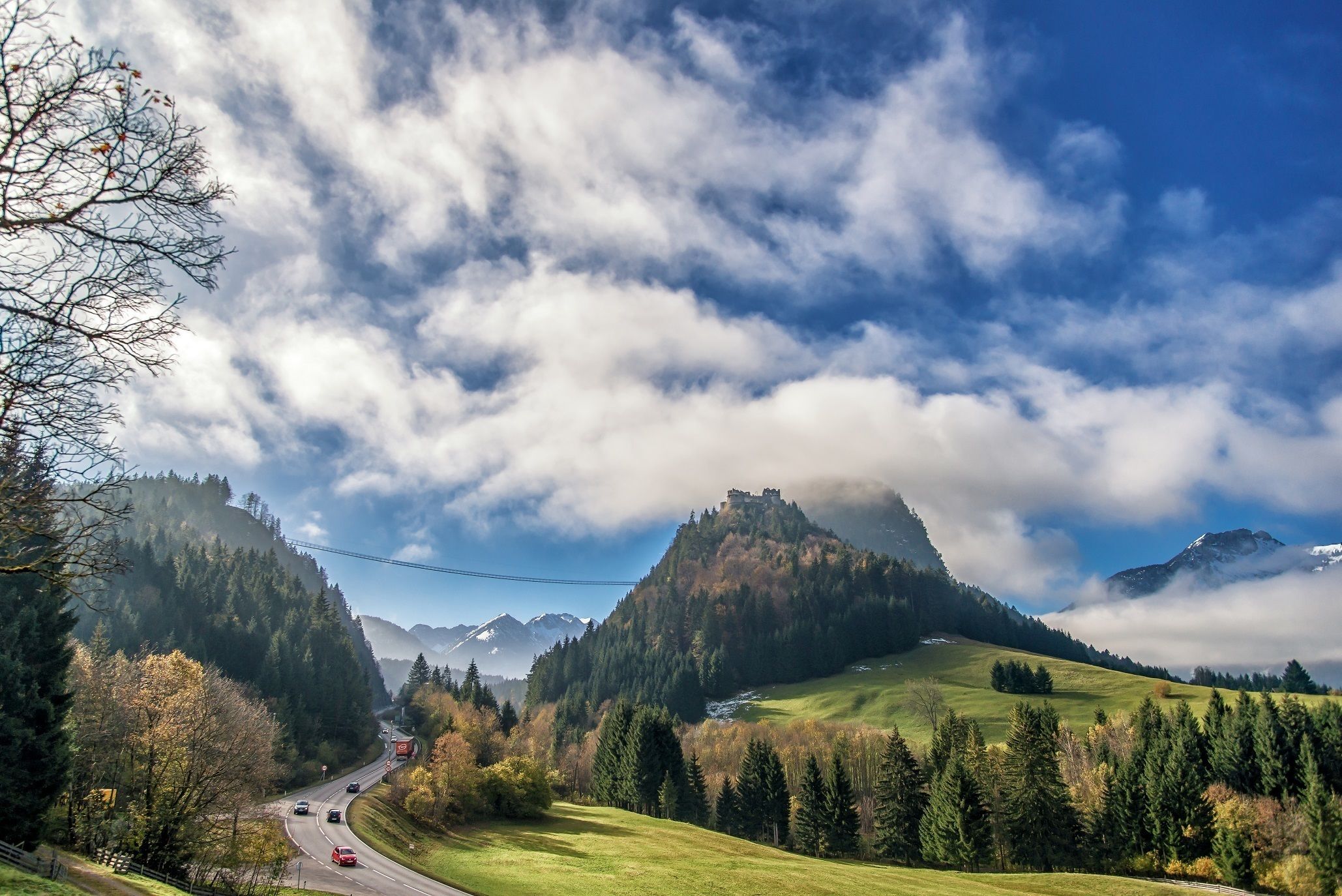 A scenic landscape with a castle on a hill, a road with vehicles, and a forested valley.