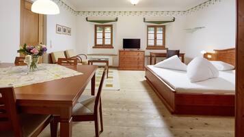 Ein Zimmer mit Esstisch aus Holz, Stühlen, einem Bett mit weißer Bettwäsche, einem Fernseher und einer Kommode.