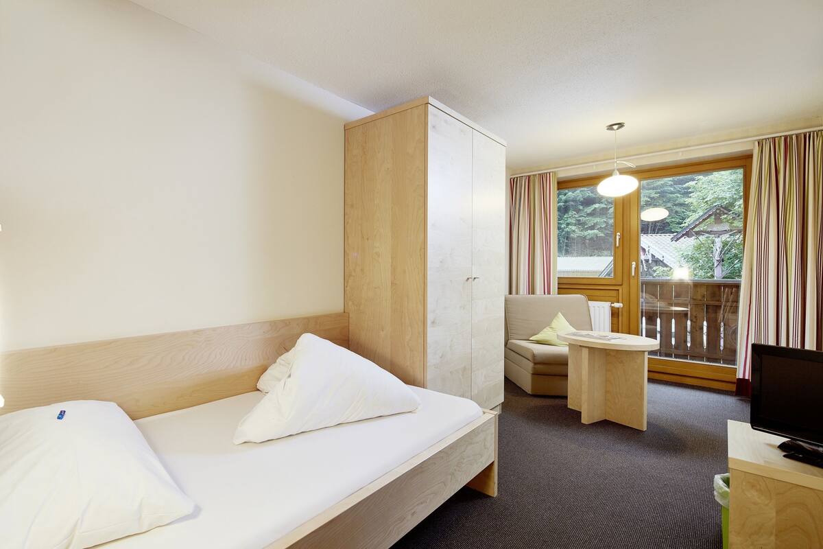 Hotellihuone, jossa on sänky, puinen vaatekaappi, pieni pöytä, televisio ja parveke, jossa on verhot.