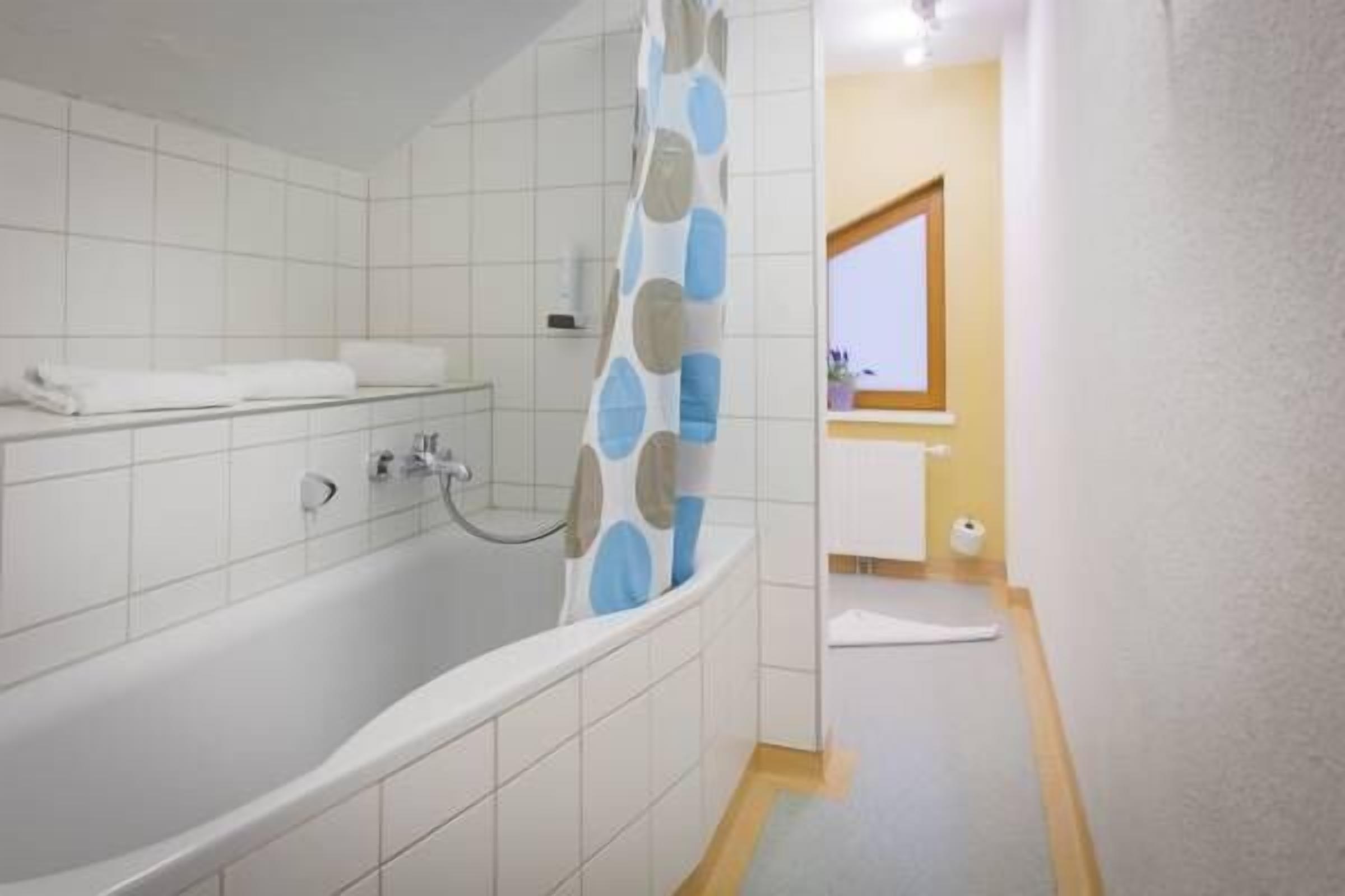 Un baño con bañera, azulejos blancos, una cortina de ducha con lunares azules y grises, y un estante pequeño con artículos de tocador.