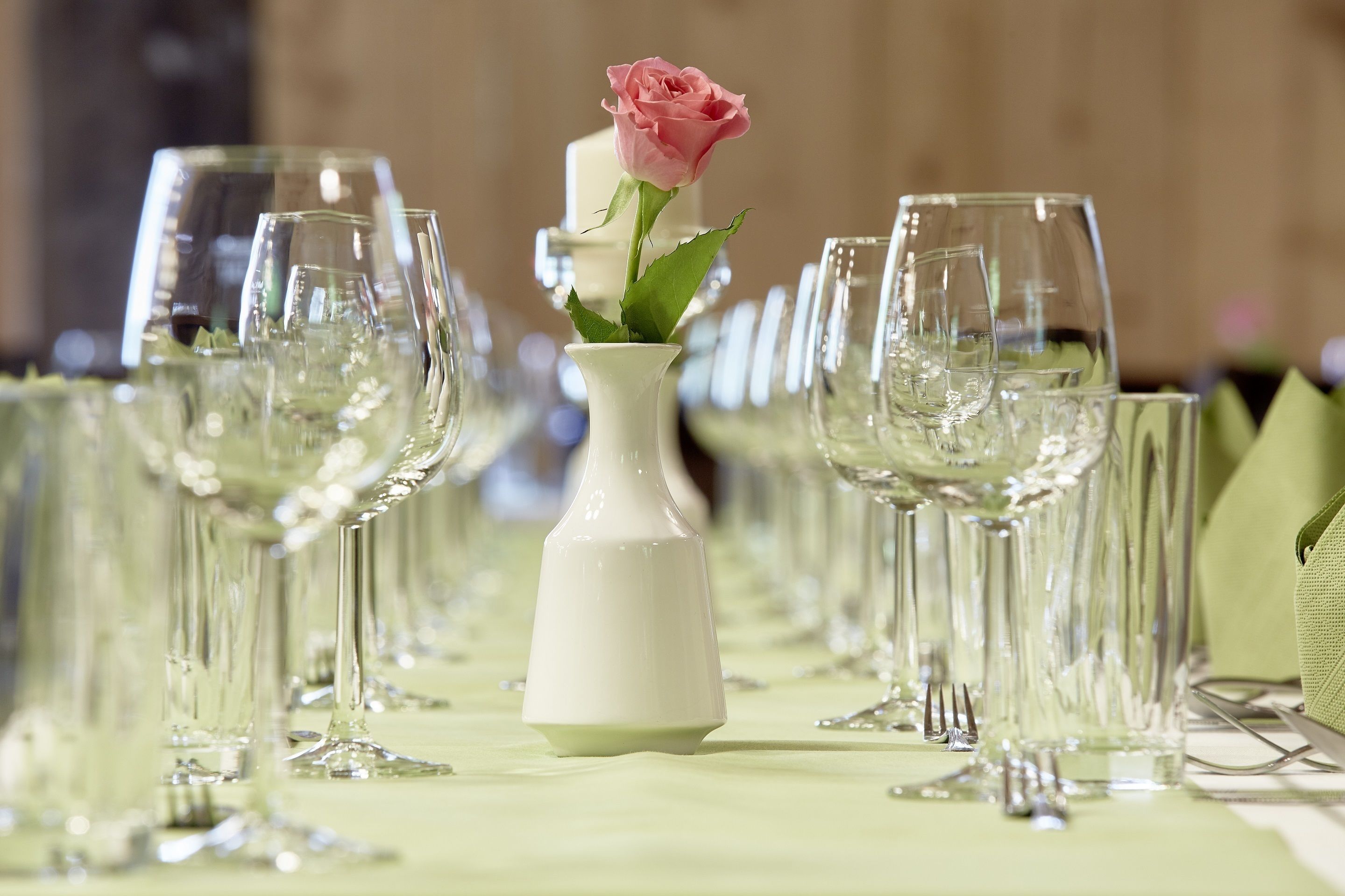 Une table dressée avec plusieurs verres à vin et un vase blanc contenant une rose rose.