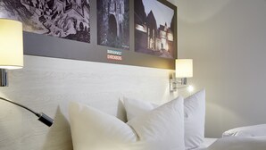 Habitación de hotel con cabecera de madera, dos lámparas fijadas a la pared y cama con ropa de cama blanca.