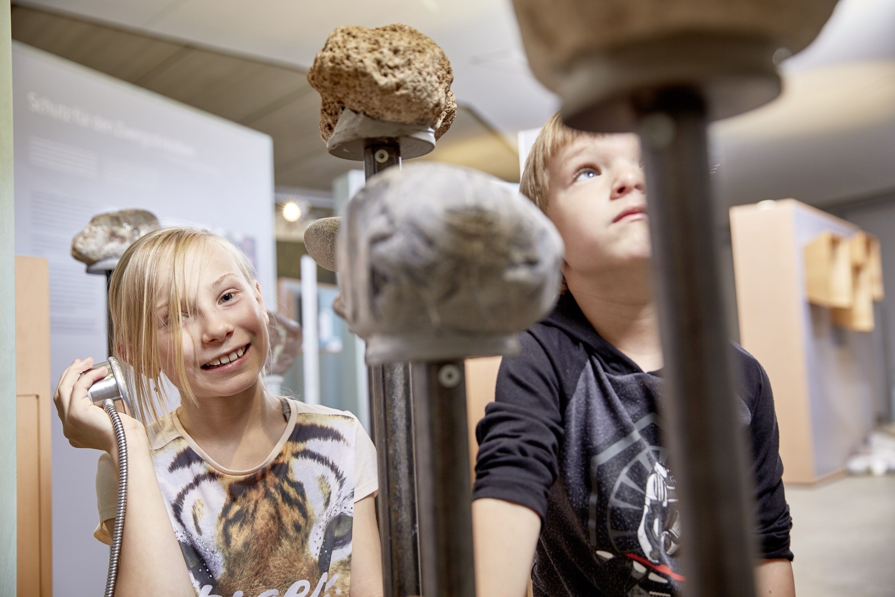 A child holding a toy telephone to a child's ear, with a display of rocks and a wall with text in the background.