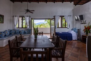 A room with a wooden table and chairs, a sofa with patterned cushions, a bed with a blue cover, and a TV on the wall. - Villas Mymosa (Quepos)