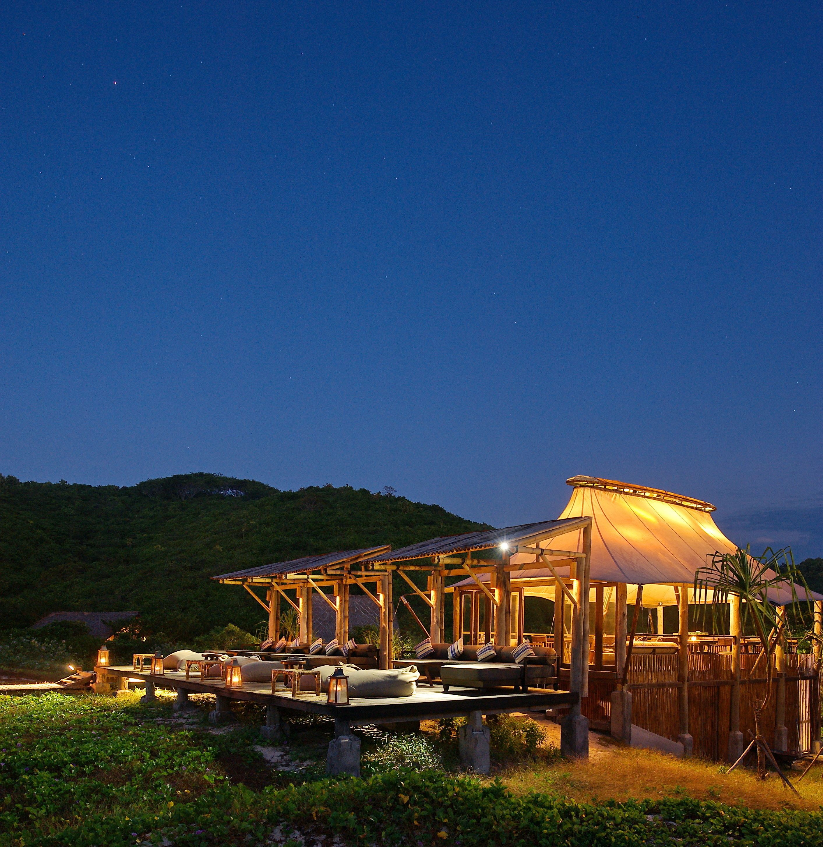 A wooden structure with a covered seating area, surrounded by greenery and hills in the background.