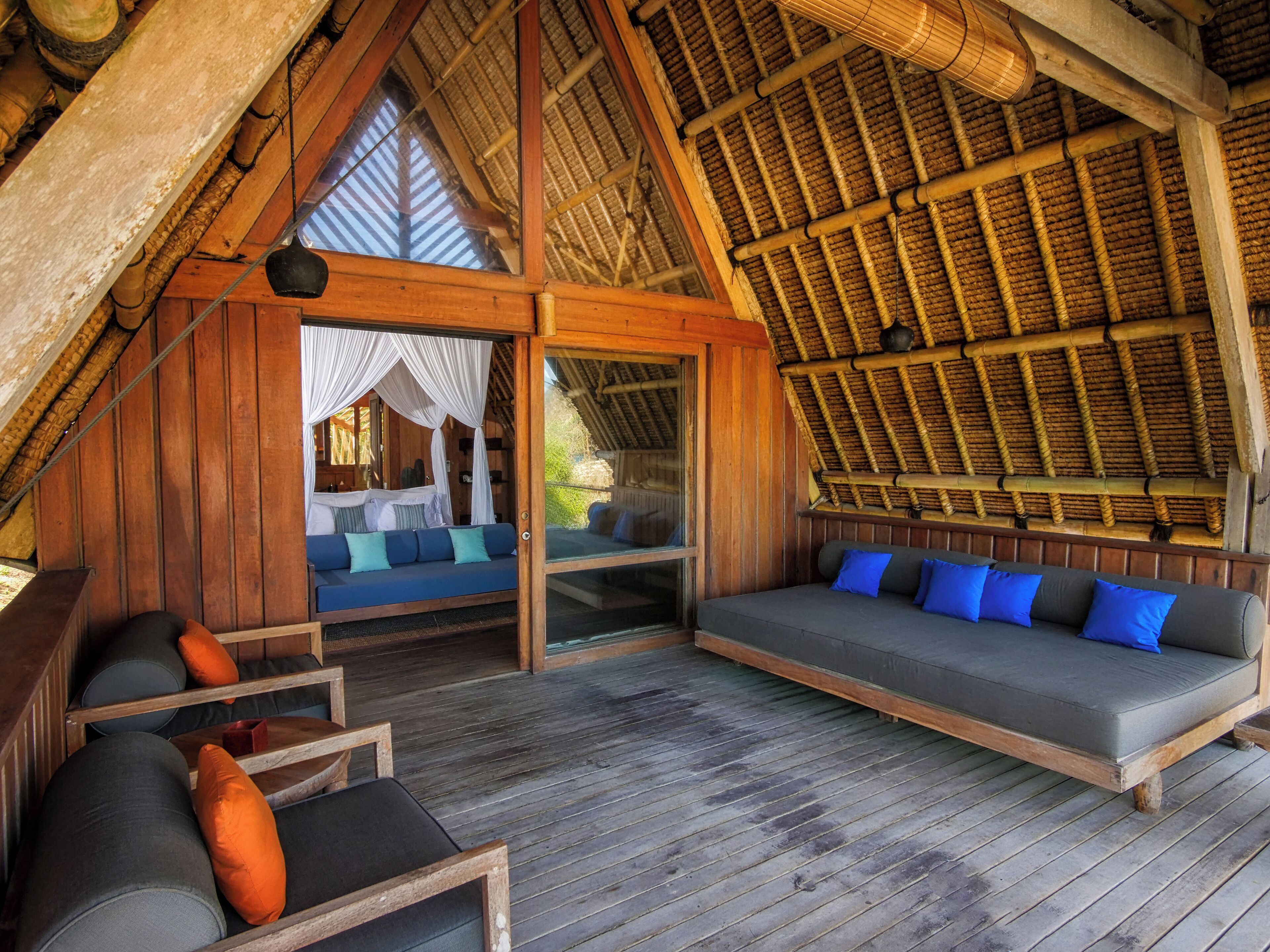 Uma cabana acolhedora de madeira com telhado de palha, um sofá grande com almofadas azuis e uma cama com cortinas brancas.