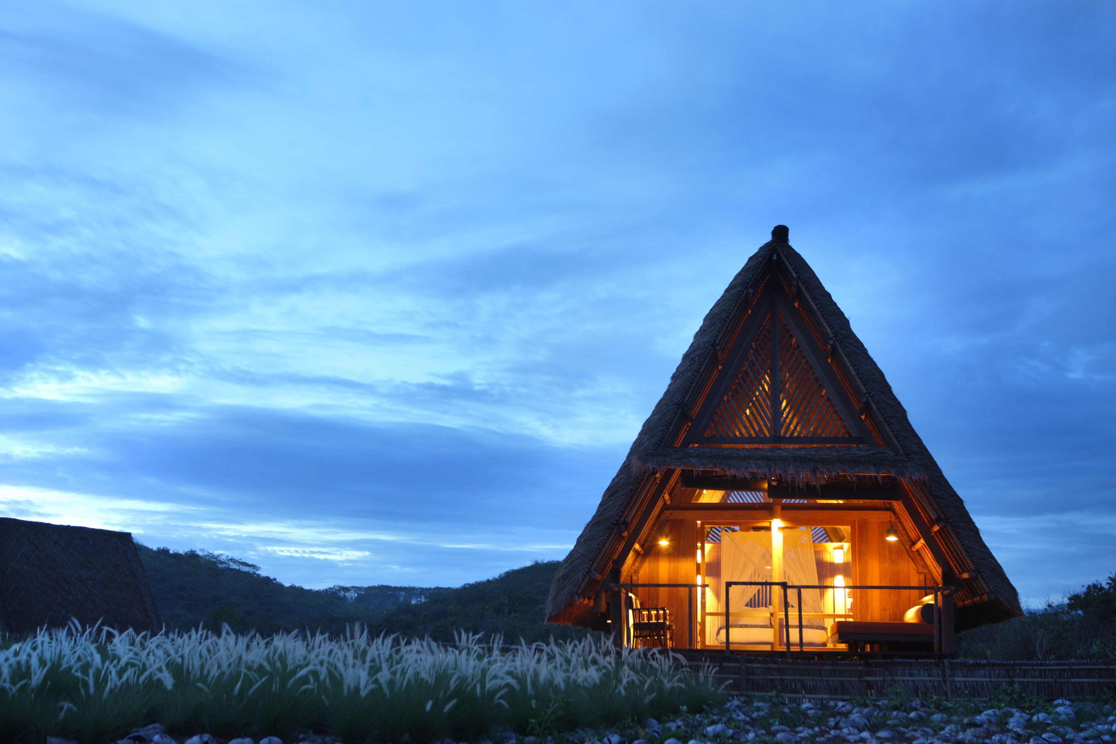 Un bâtiment triangulaire à toit de chaume, doté d’un intérieur ouvert et entouré d’herbes hautes et de collines.