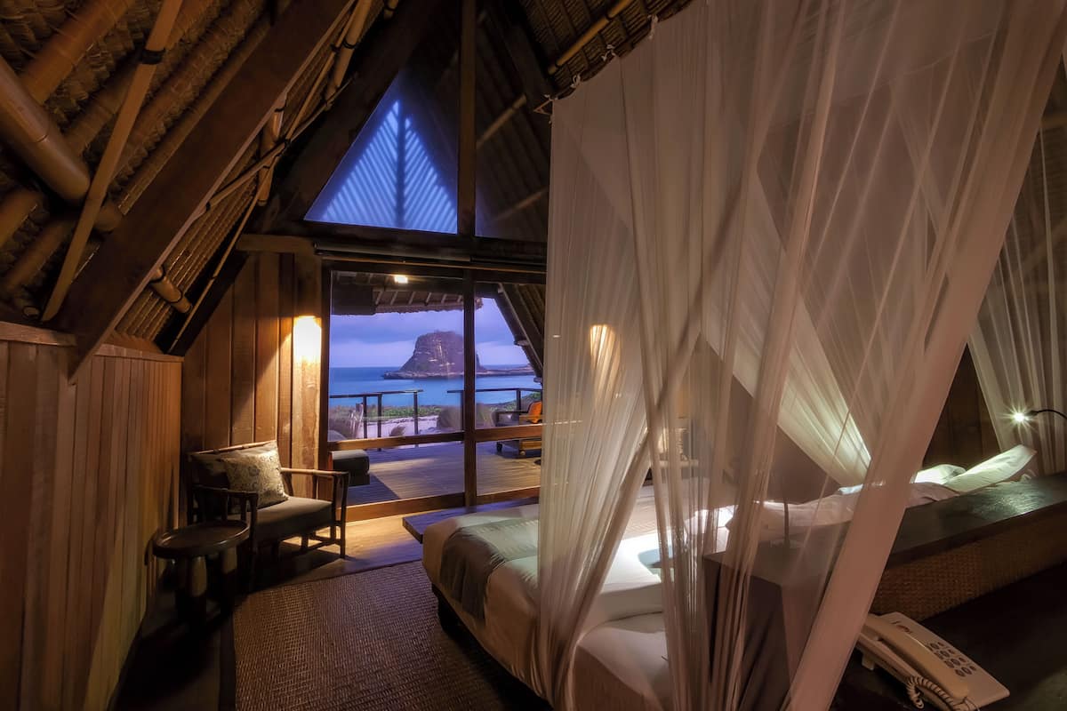 A room with a bed, a table, and a chair. There is a view of the sea and a mountain through a window.