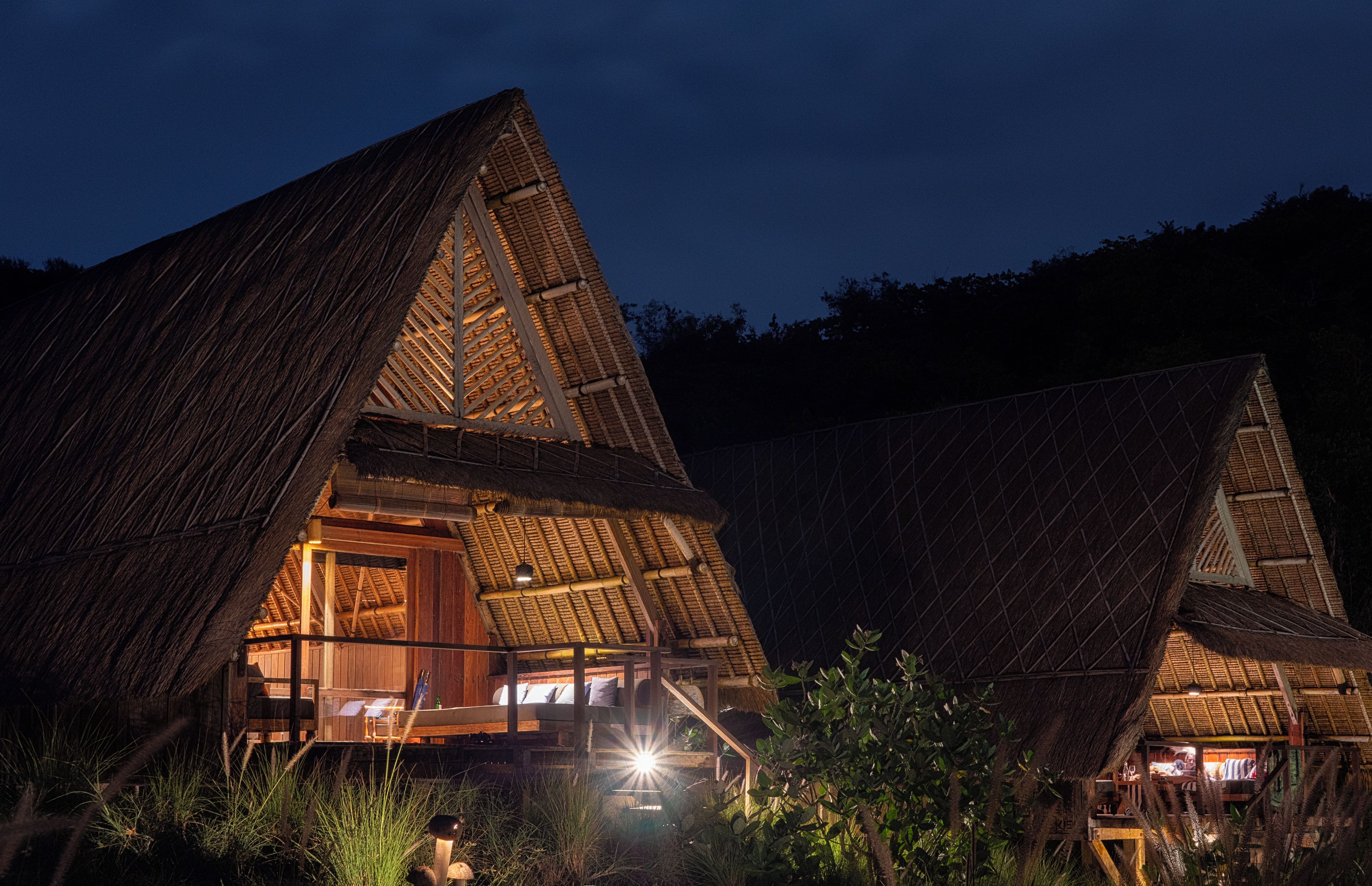 Two thatched-roof cottages with wooden interiors, surrounded by grass and trees, at night.