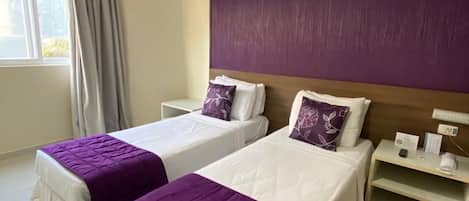 Una habitación de hotel con dos camas, una mesita de noche, una ventana con cortinas y una rejilla en el techo.