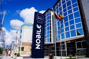 Un hotel chiamato 'Noble Suites' con una facciata moderna in vetro, un cartello ben visibile e un ristorante chiamato 'Kura'.