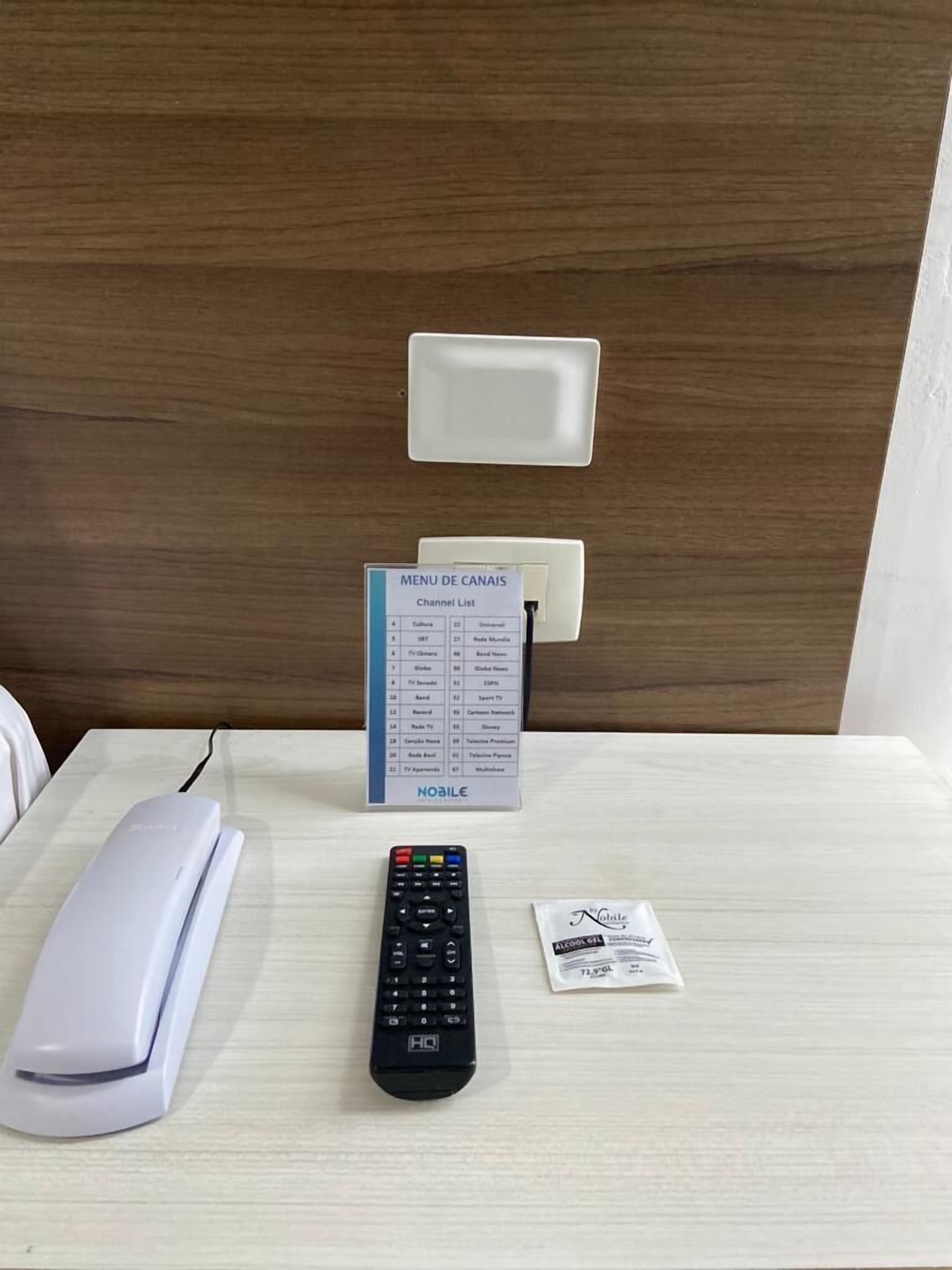 Une chambre d’hôtel avec une table de chevet blanche, une télécommande, un téléphone portable, un panneau indiquant la liste des chaînes et un mur blanc.
