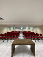 A conference room with rows of red chairs, a wooden table, and a white tiled floor.
