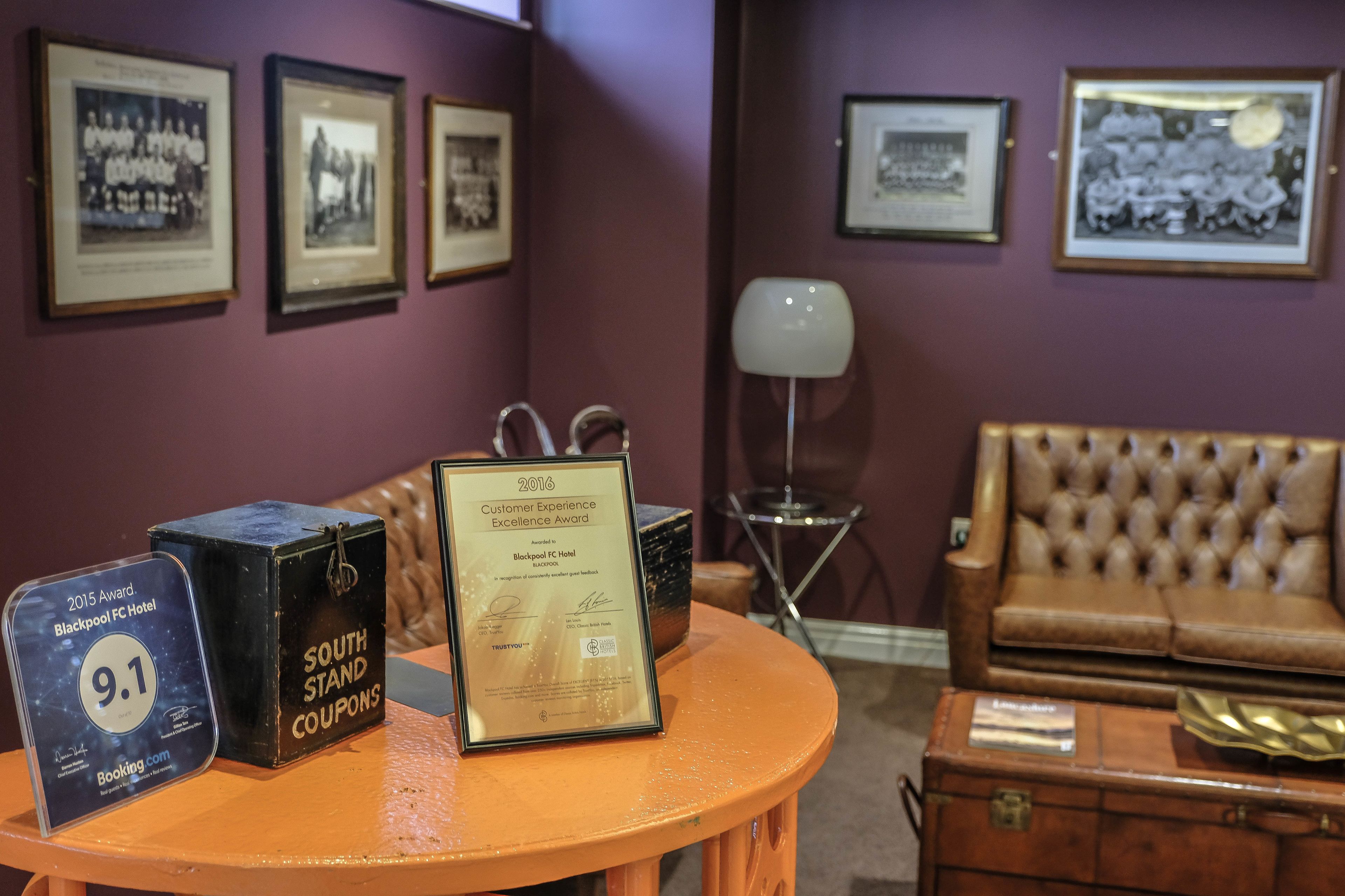 A room with a round table displaying awards and a framed certificate, a tufted leather sofa, and framed pictures on the wall.