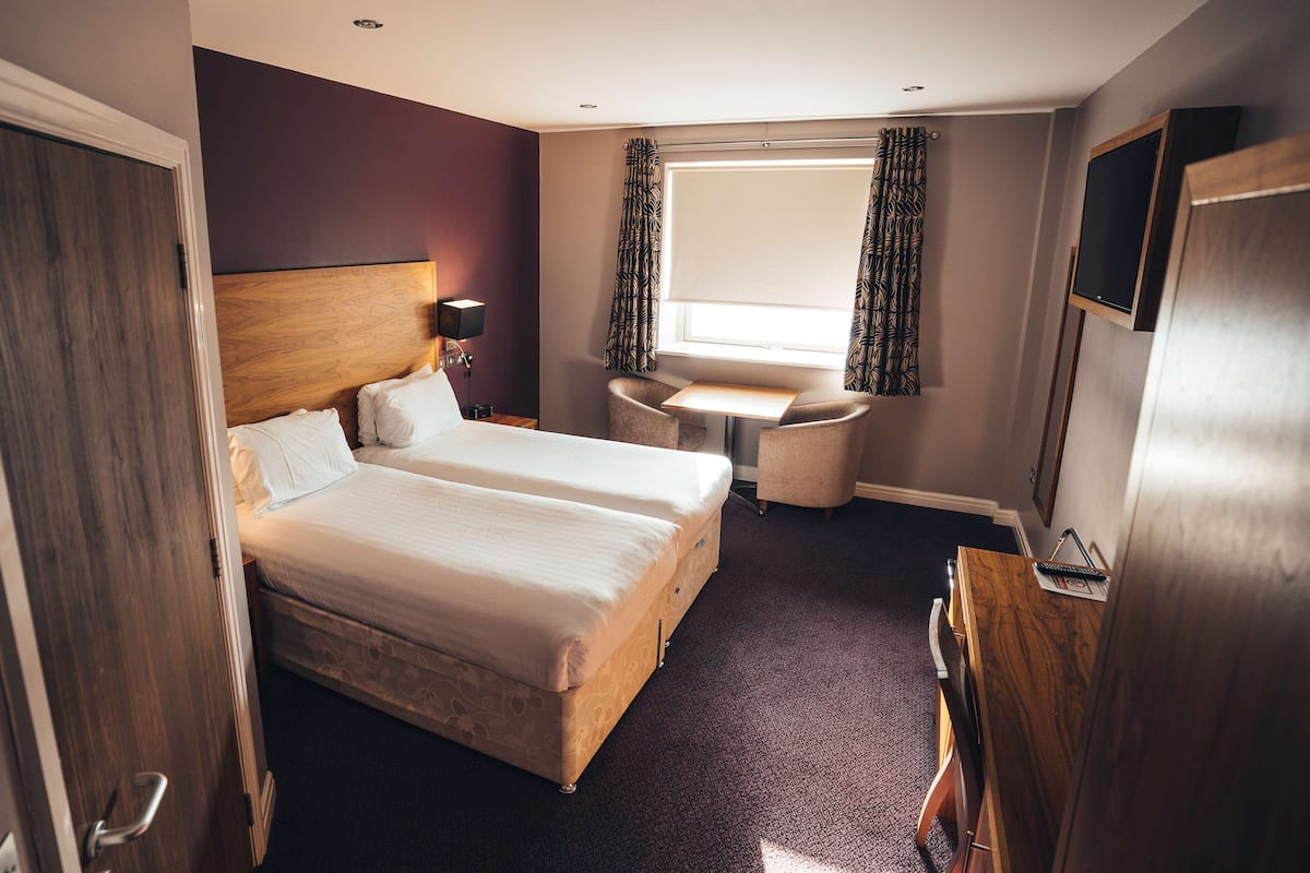 A hotel room with two beds, a desk, a chair, a TV, and a window with curtains.