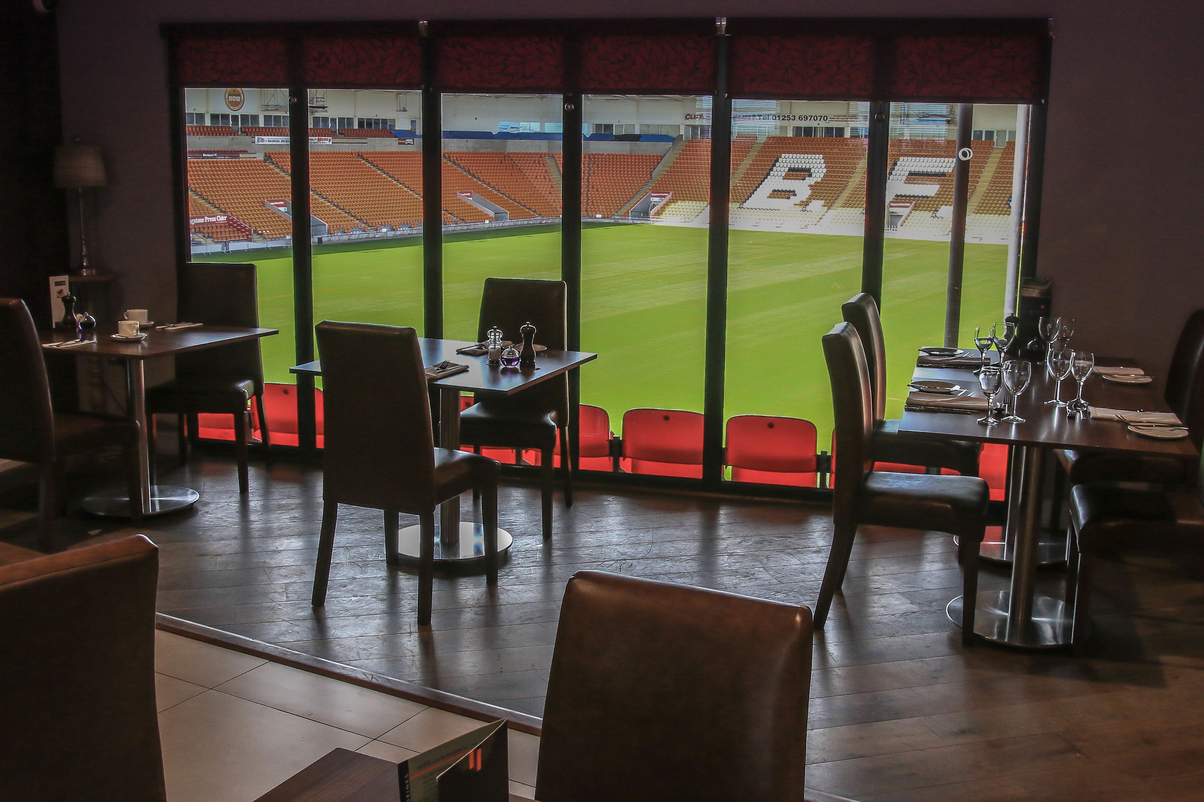 Un restaurant avec vue sur un stade. Les tables sont disposées avec des chaises, et on y trouve des verres à vin et des tasses.