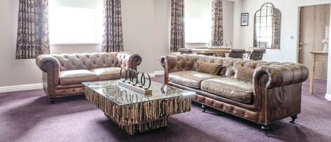 A living room with two leather sofas, a glass coffee table, and large windows with patterned curtains.