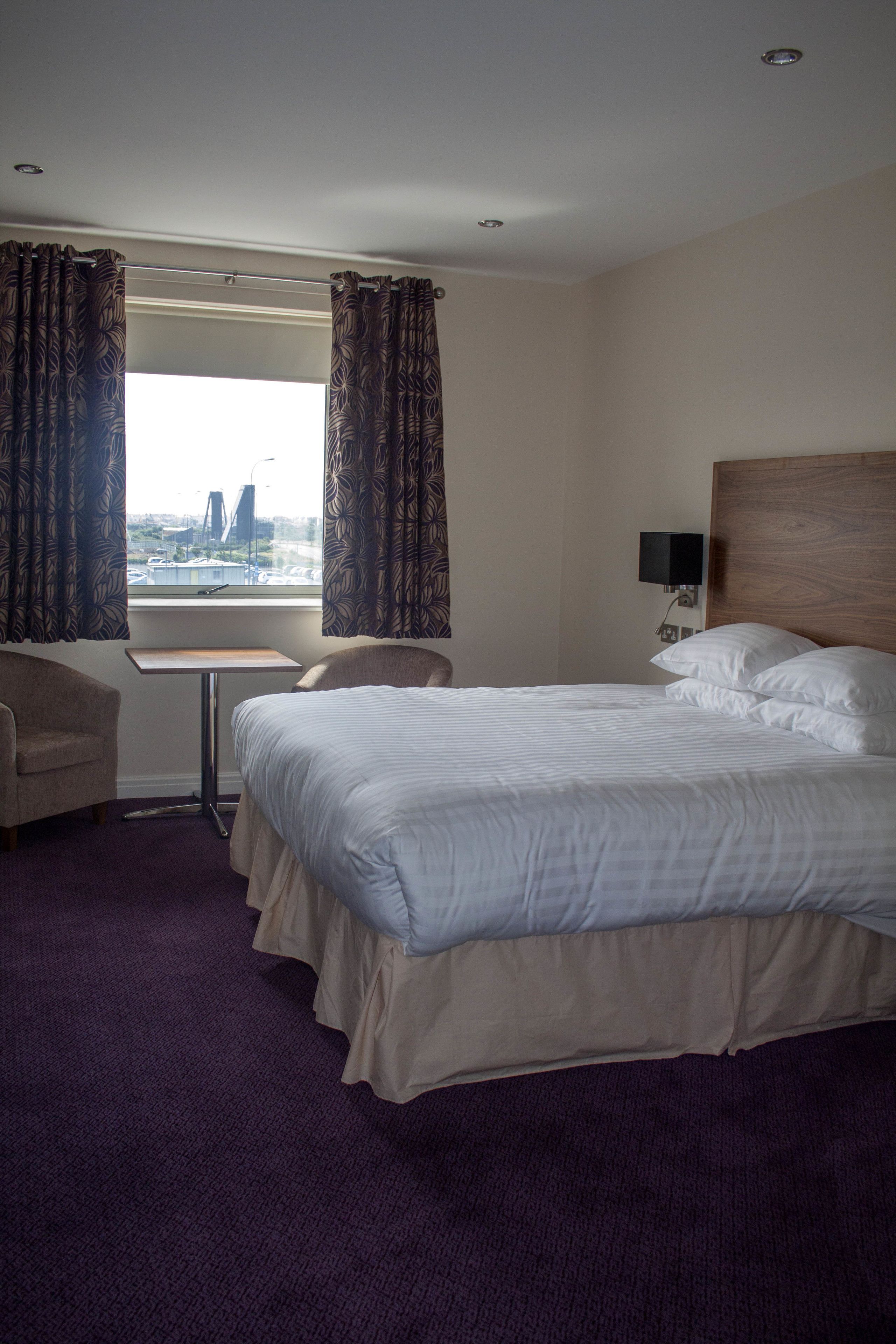 Habitación de hotel con una cama grande, un escritorio y una silla. Tiene una ventana con cortinas, vistas a un puente y un televisor montado en la pared.