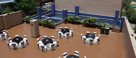 A rooftop terrace with multiple round tables and white chairs, surrounded by potted plants and a swimming pool.