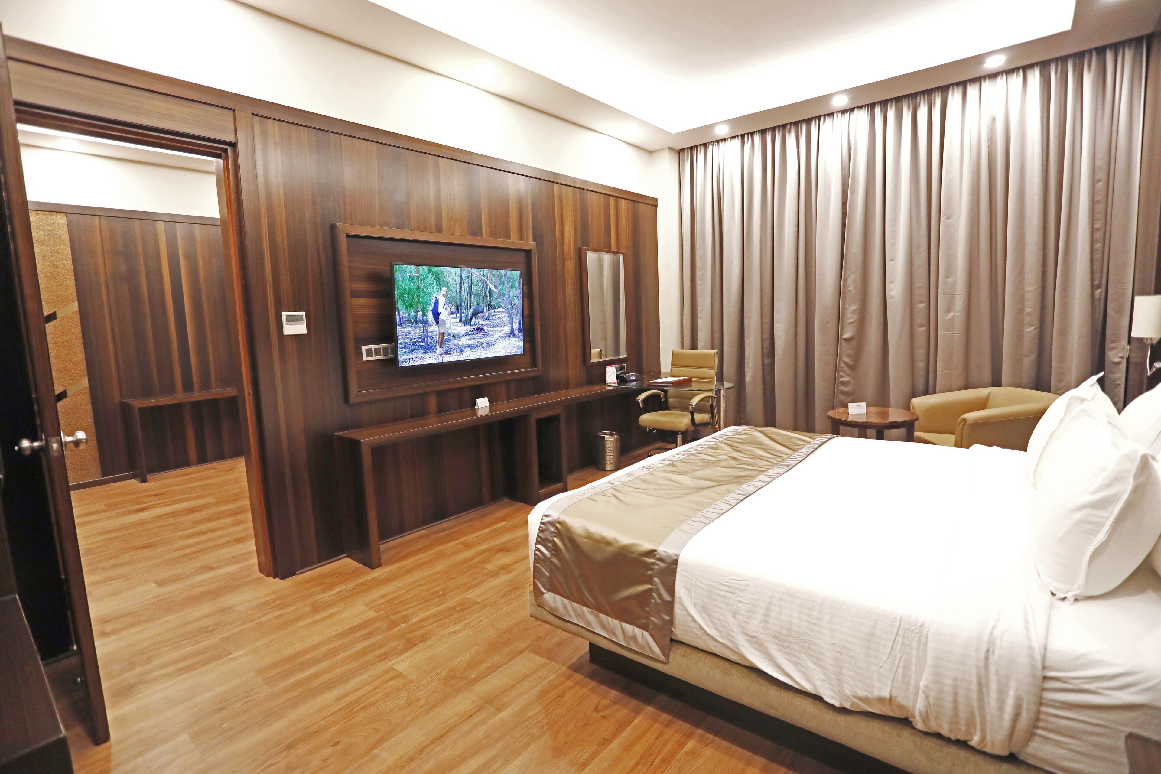 A hotel room with a large bed, a television mounted on the wall, a seating area with a chair and a small table, and a wardrobe.