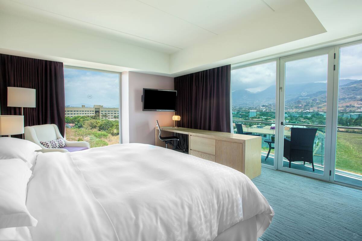 A hotel room with a large bed, a desk with a chair, a TV, and a balcony with a view of the city and mountains.