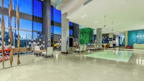 A spacious lobby with large windows, a green wall feature, and a seating area with tables and chairs.