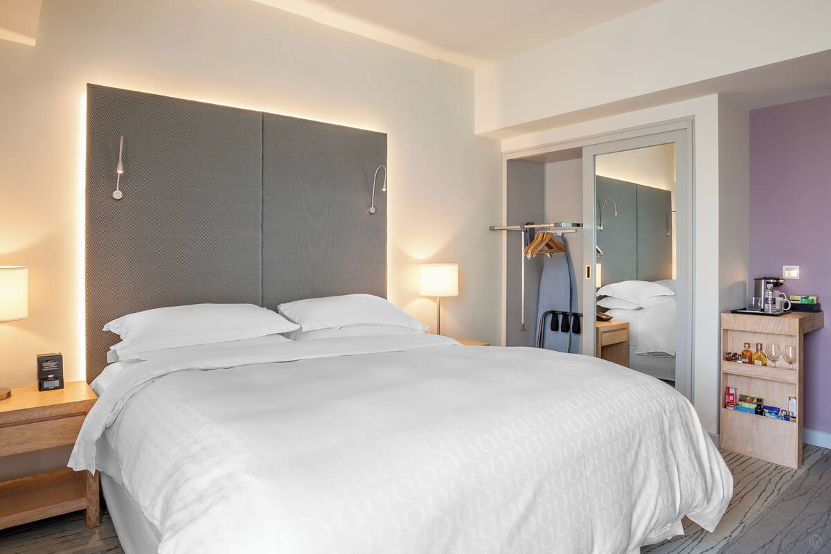 A hotel room with a large bed, bedside lamps, a wooden nightstand, a mirror, and a small wooden shelf with toiletries.