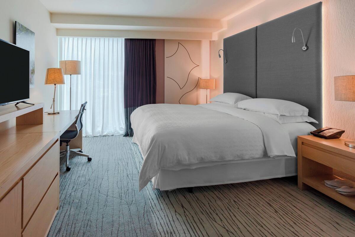 A hotel room with a bed, bedside tables, a desk, a chair, a TV, and a window with curtains.