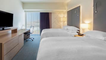 A hotel room with two beds, a desk with a TV, a chair, and a balcony with curtains.