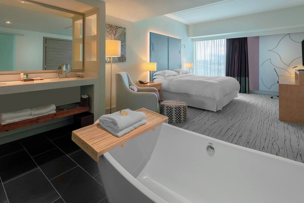 A modern hotel room with a large bathtub, a separate bedroom with a bed, a sitting area with a chair, a vanity with a mirror, and a large window with curtains.