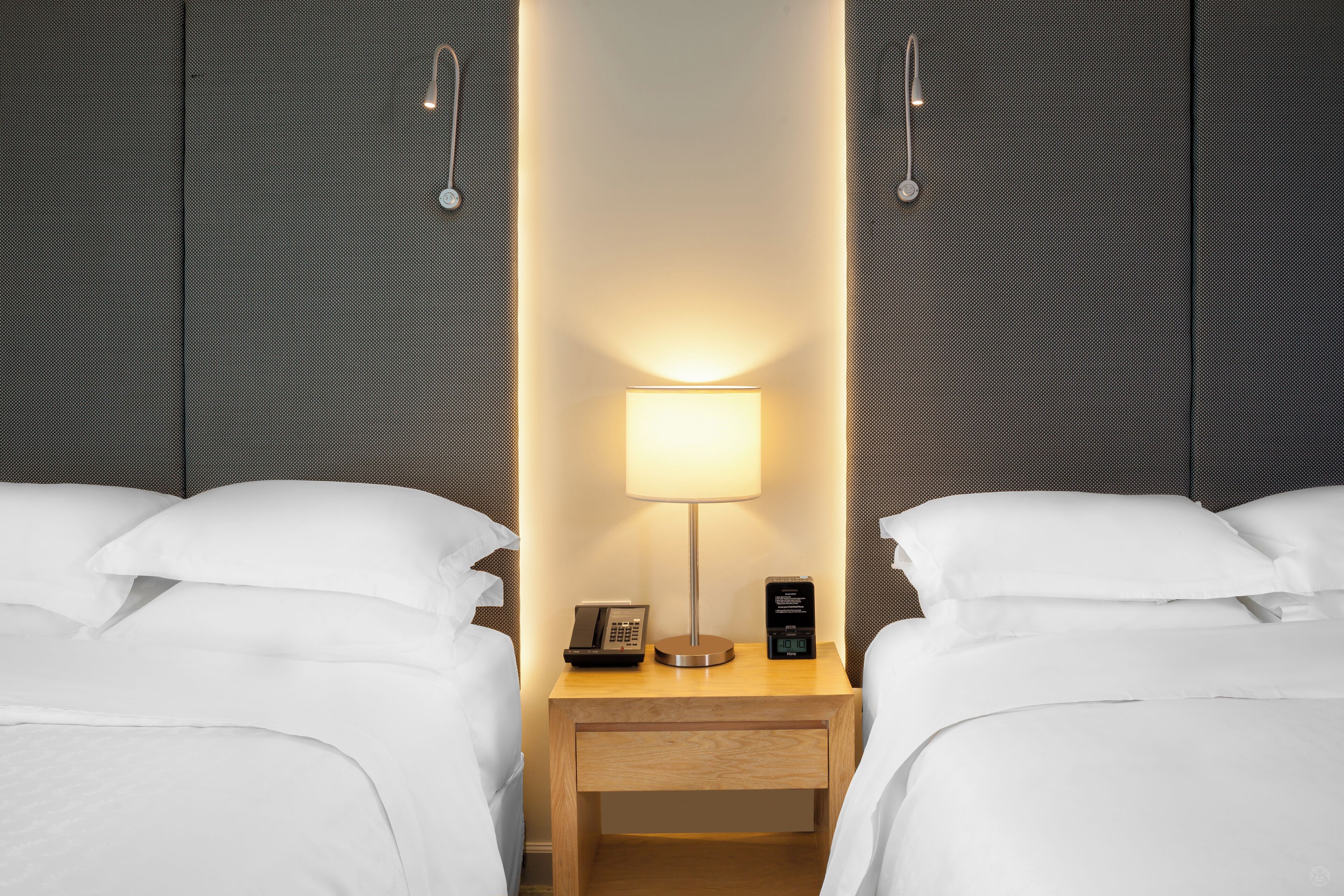 Une chambre d’hôtel avec deux lits, une table de chevet avec une lampe, un téléphone et une horloge numérique.