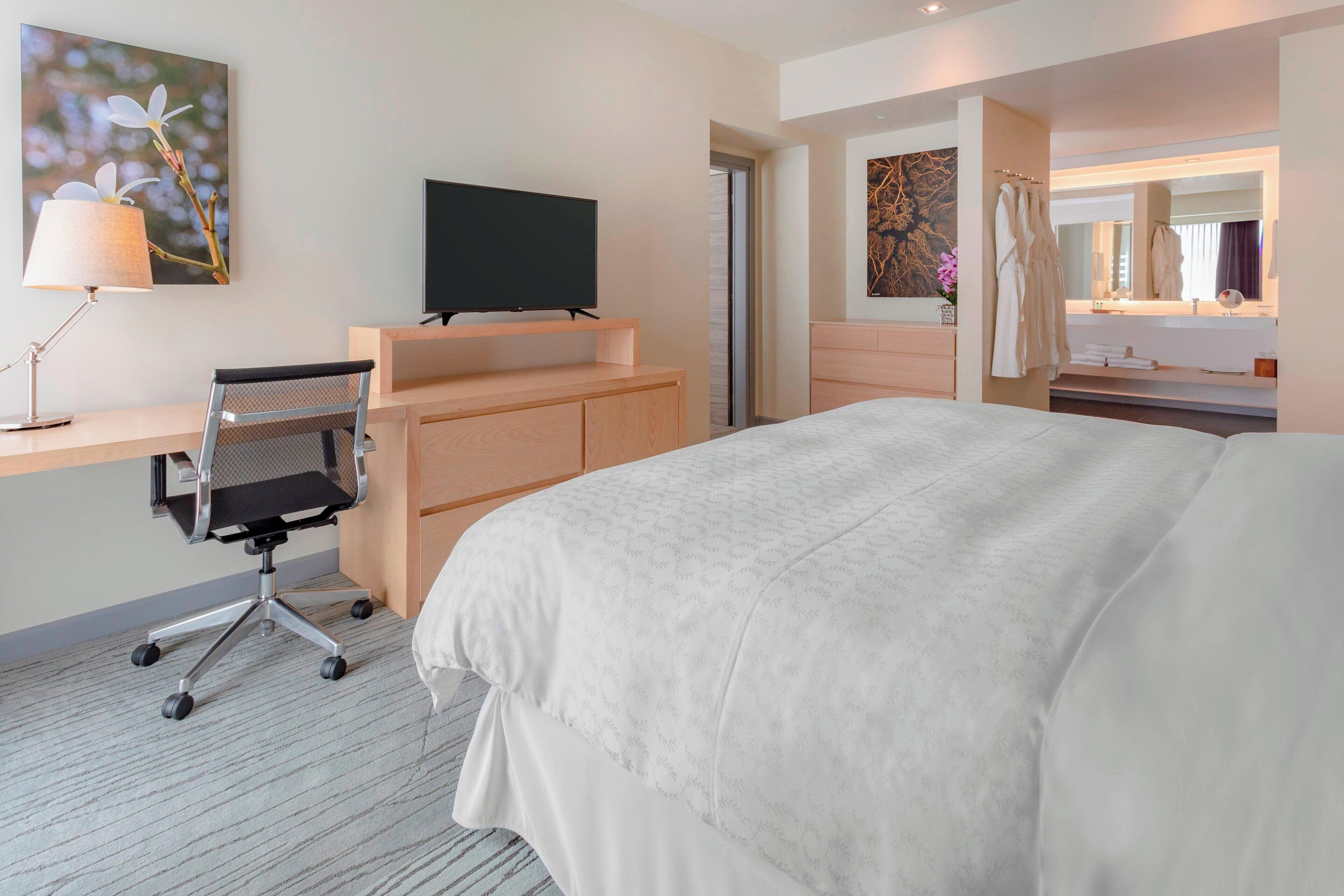 A hotel room with a large bed, a desk with a chair, a flat-screen TV, a bathroom with a mirror, and a painting on the wall.