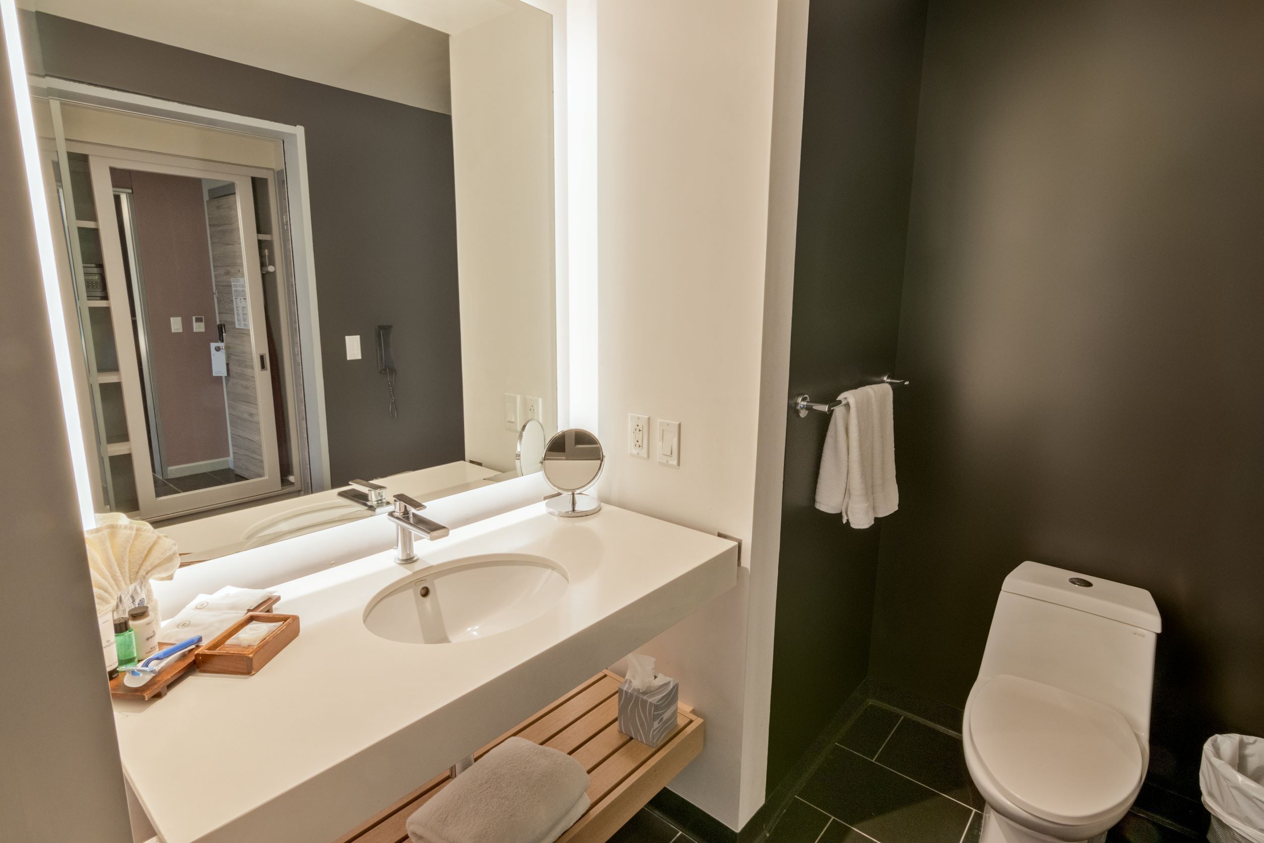 Un baño moderno con un lavabo blanco, espejo e inodoro. Hay un estante de madera debajo del lavabo y una toalla colgada en la pared.